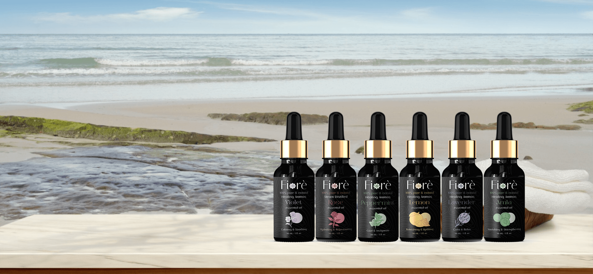 Six Fiore essential oil bottles displayed on a wooden surface by the beach, featuring Violet, Rose, Peppermint, Lemon, Lavender, and Amla oils.